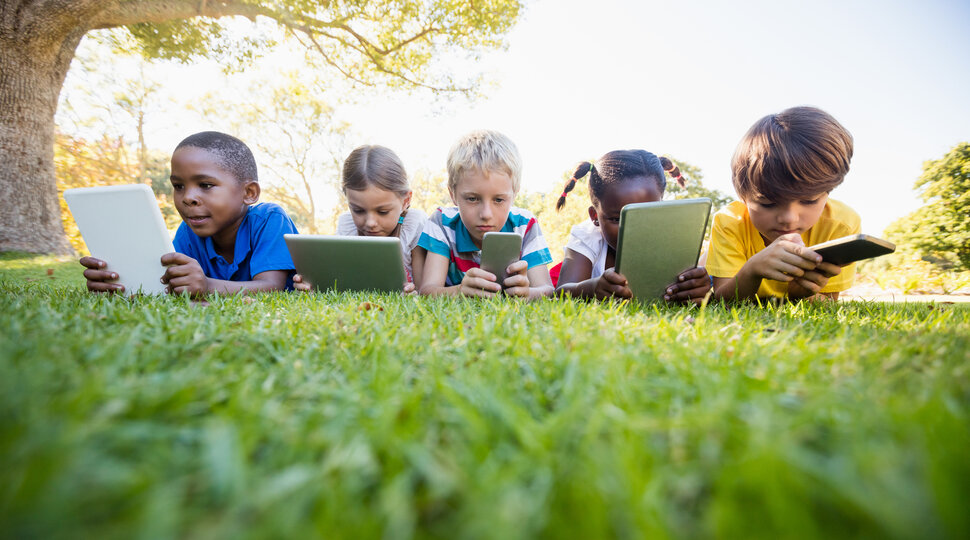Fünf Kinder, die an einem sonnigen Tag mit digitalen Geräten im Park liegen