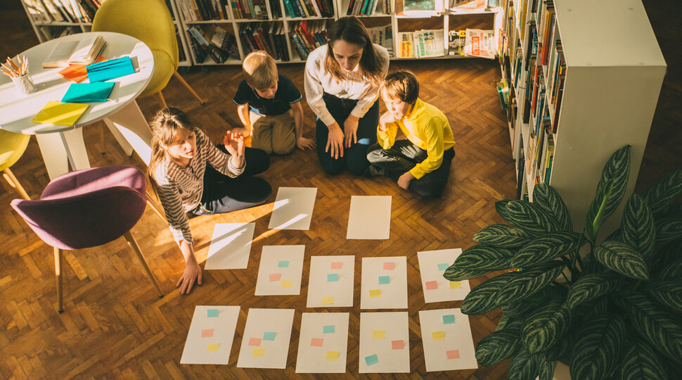 Kinder und Jugendliche sitzen mit vielen Zetteln und Post-its auf dem Fußboden einer Bibliothek