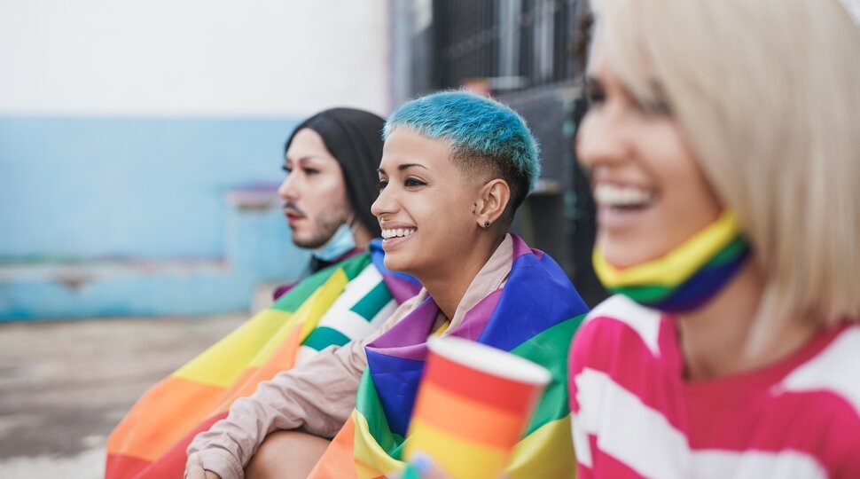 Drei queere mit Regenbogenfahnen lachen und schauen in die Ferne