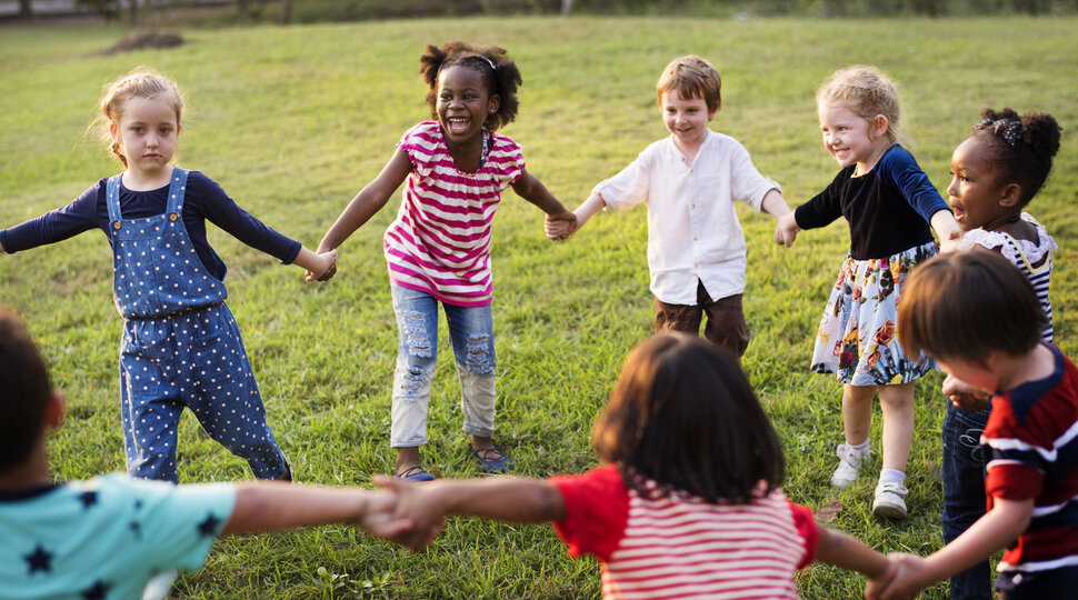 Verschiedene Kinder stehen auf einer Wiese im Kreis und halten sich an den Händen