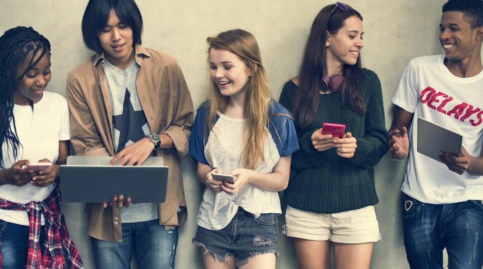Fünf Jugendliche stehen an einer Wand, schauen auf Bildschirme und lachen