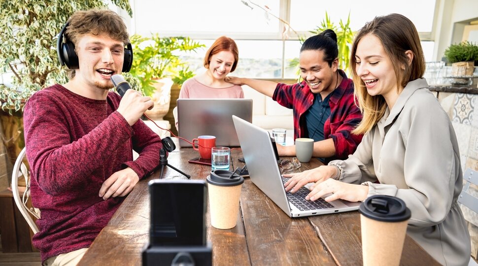 Vier Jugendliche sitzen an einem Tisch an zwei Laptops und und nehmen ein Video mit dem Smartphone auf