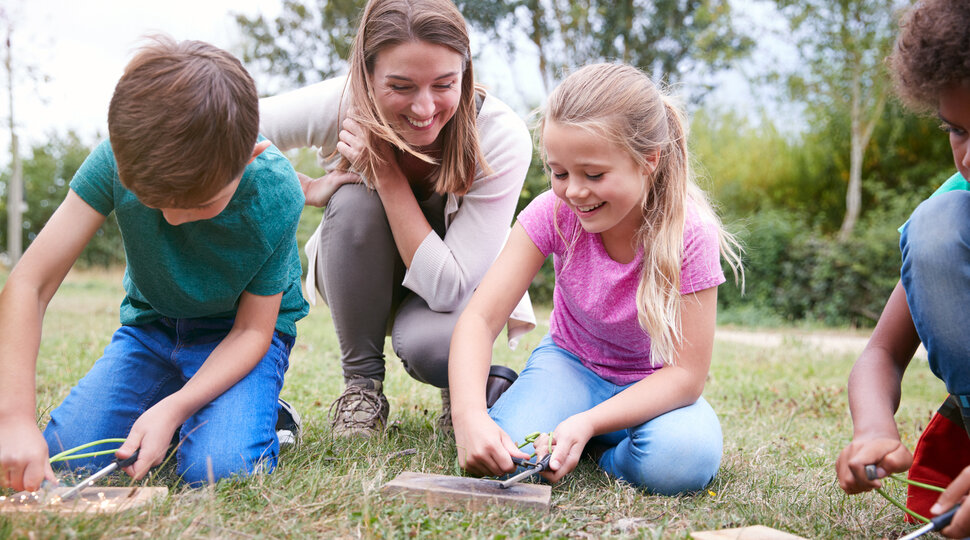 Drei Kinder sitzen mit einer Erwachsenen auf einer Wiese und bauen etwas