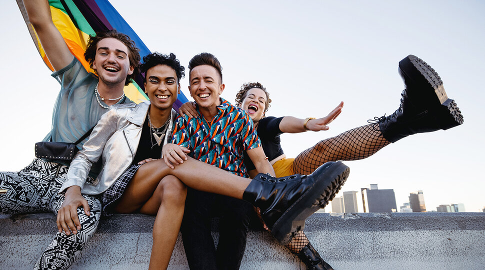 Vier LGBTQ-Freunde feiern und lachen, während sie eine Regenbogenflagge in die Luft halten