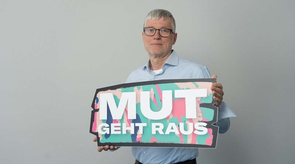 Ein Mann mit grauen Haaren und einer Brille hält ein Schild in der Hand mit der Aufschrift: "Mut geht raus"