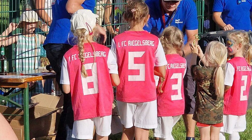 fünf Mädchen in rosa Trikots mit weißen Hosen bekommen von Männern in blauen T-Shirts einen goldenen Pokal überreicht