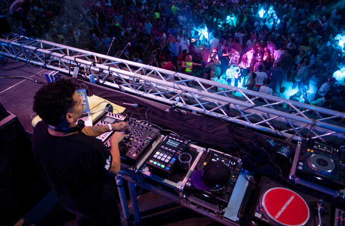 DJ steht am Pult, von hinten zu sehen und blickt in eine tanzende Menge, beleuchtet von blau und pinkfarbenem Neonlicht