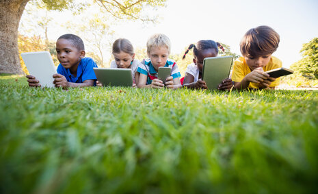 Fünf Kinder, die an einem sonnigen Tag mit digitalen Geräten im Park liegen
