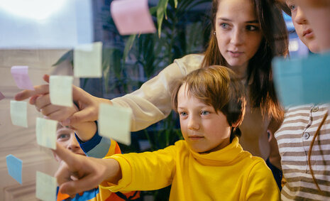 Kinder und Jugendliche zeigen auf bunte Post-its, die am Fenster hängen