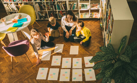 Kinder und Jugendliche sitzen mit vielen Zetteln und Post-its auf dem Fußboden einer Bibliothek
