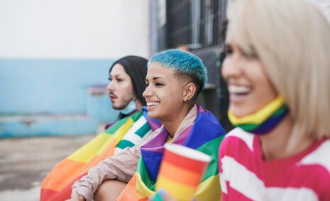 Drei queere mit Regenbogenfahnen lachen und schauen in die Ferne