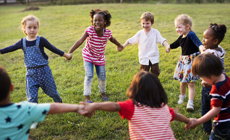 Verschiedene Kinder stehen auf einer Wiese im Kreis und halten sich an den Händen