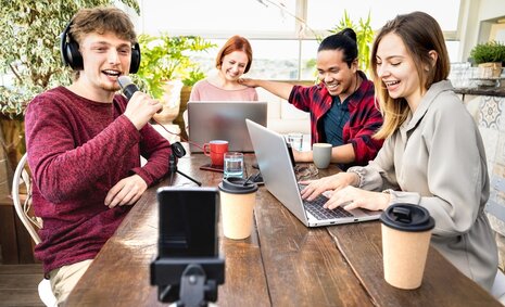 Vier Jugendliche sitzen an einem Tisch an zwei Laptops und und nehmen ein Video mit dem Smartphone auf