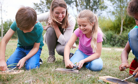 Drei Kinder sitzen mit einer Erwachsenen auf einer Wiese und bauen etwas