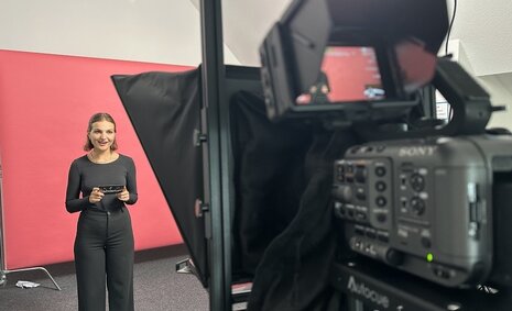 Eine junge Frau steht im Hintergrund links vor einem orangefarbenen Hintergrund, im Vordergrund steht eine Kamera, die Frau hält Postkarten in der Hand.