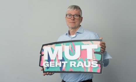 Ein Mann mit grauen Haaren und einer Brille hält ein Schild in der Hand mit der Aufschrift: "Mut geht raus"